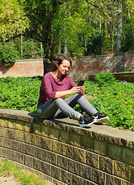 Marlies zit op een muurtje in een park met een mobiele telefoon in de hand.